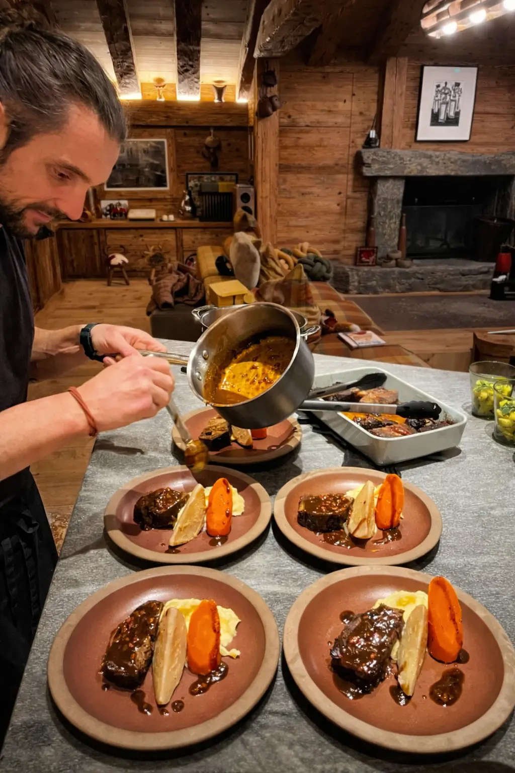 Man with a beard and tied-back hair plating brown sauce from a pot onto four dishes with meat, mashed potatoes, and vegetables in a rustic wooden kitchen.