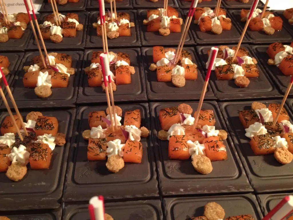 Rows of bite-sized appetizers with diced orange pieces, white cream dollops, herbs, and small round crackers on black square plates with toothpicks.