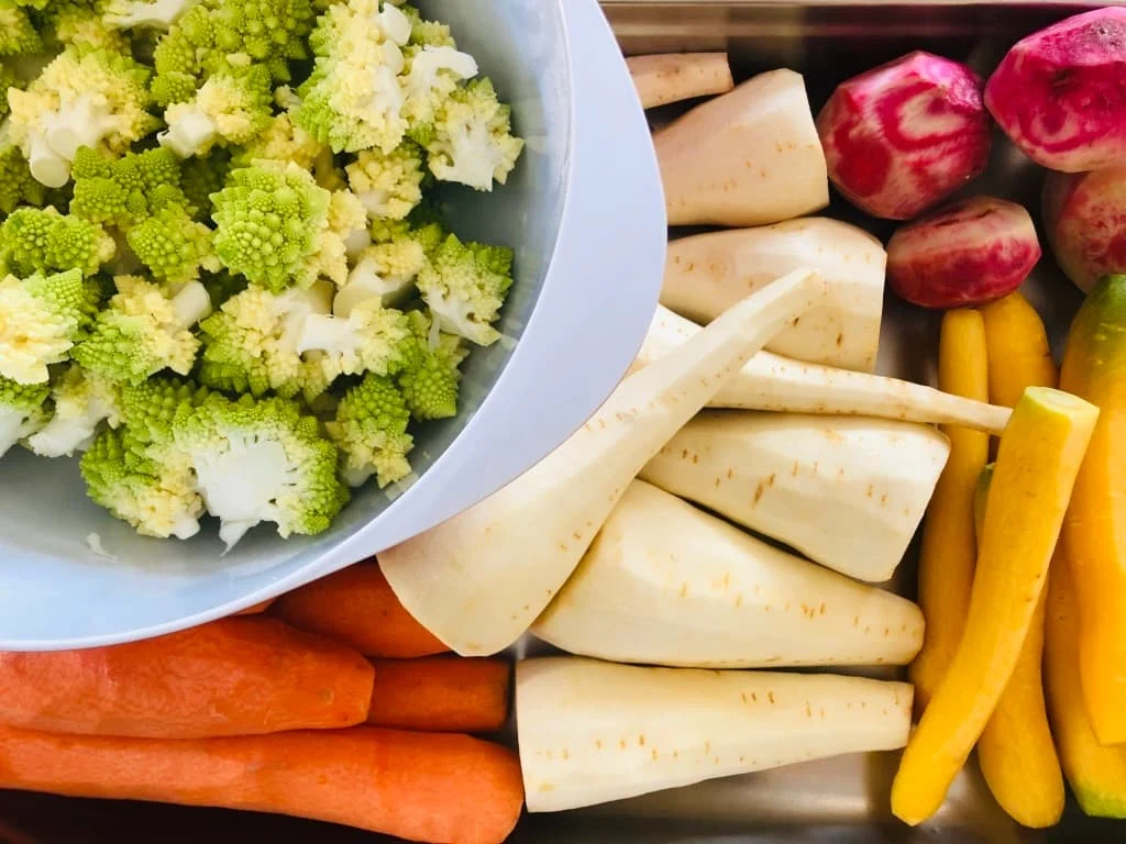 Various fresh vegetables including green Romanesco broccoli florets in a bowl, peeled parsnips, whole carrots, striped beets, and yellow squash.