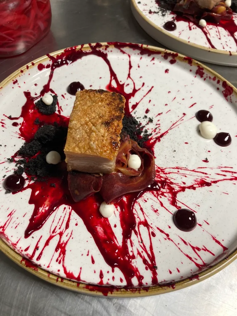 Plate with seared pork belly on beet puree, garnished with thinly sliced cured meat, black crumbs, and small dollops of white sauce.